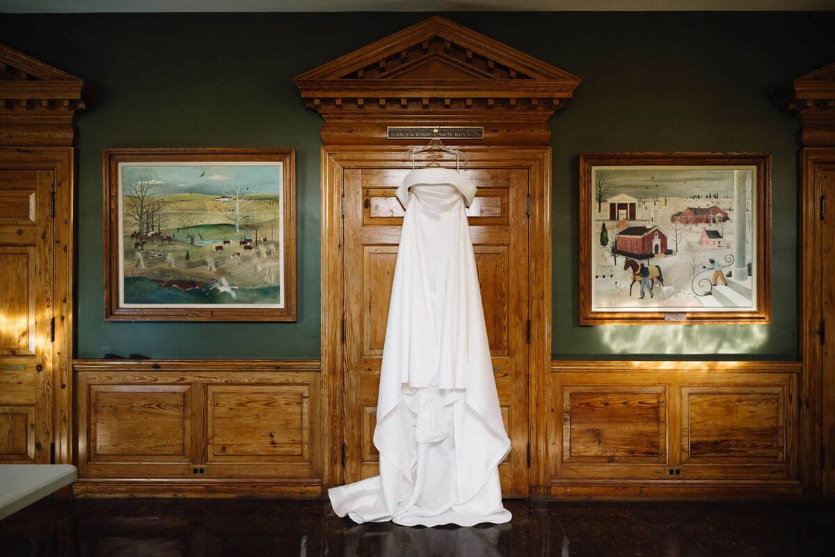 wedding photo of brides wedding dress hanging from a wooden door in a dark green room with two framed photos on either side of the door at middleburg community center