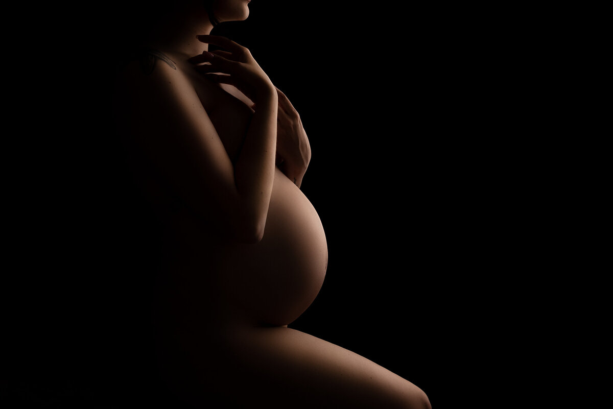 In this moody and intimate maternity photo captured by Philadelphia's premier maternity photographer Katie Marshall, an expectant mom stands side profile to the camera with her front leg bent in front of her. Her hands are fold in front of her chest and touching her chin. Her face is cropped out of the image/
