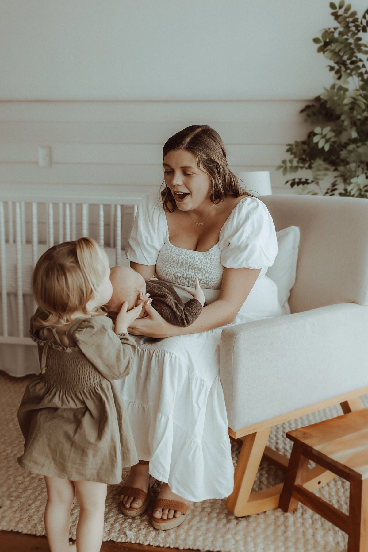 A woman in a white dress sits on a chair holding a baby, while a young child stands nearby, touching the baby. A crib and plant are in the background.