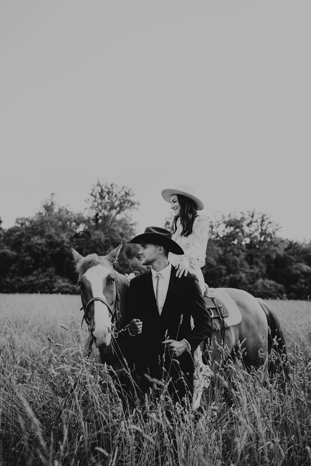 Black and white photo of a bride and groom on horseback in a tall grassy field