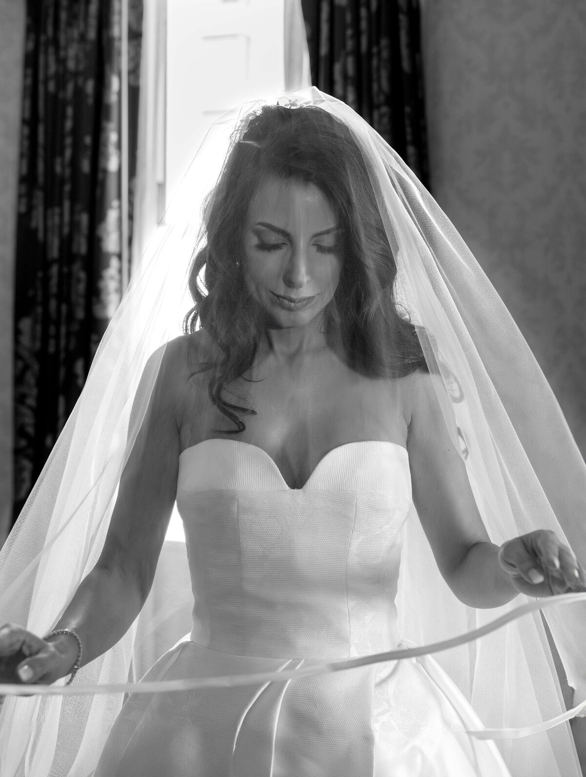 A bride in a strapless wedding gown with a veil stands by a bright window. The veil drapes over her shoulders as she gently holds onto it with both hands. The setting is elegant, featuring patterned curtains and soft lighting.