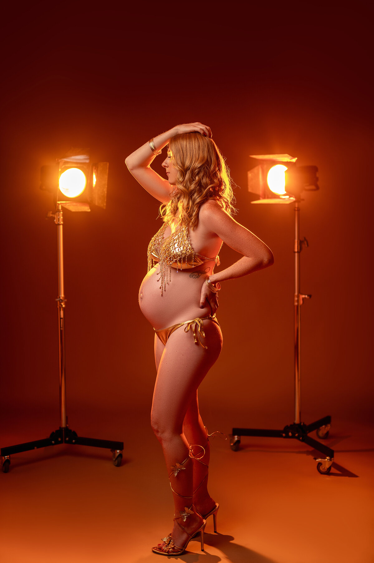 Pregnant woman posing confidently in a gold bikini and high heels, illuminated by studio lights for a bold, fashion-inspired maternity portrait in Waukesha, Wisconsin.