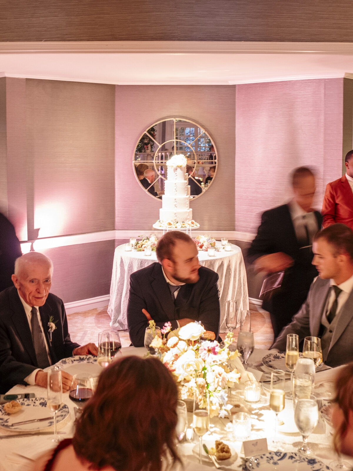 A warmly lit banquet room with guests seated at a table adorned with floral centerpieces and glasses. In the background, a tall, elegant wedding cake is displayed on a decorated table in front of a round mirror.