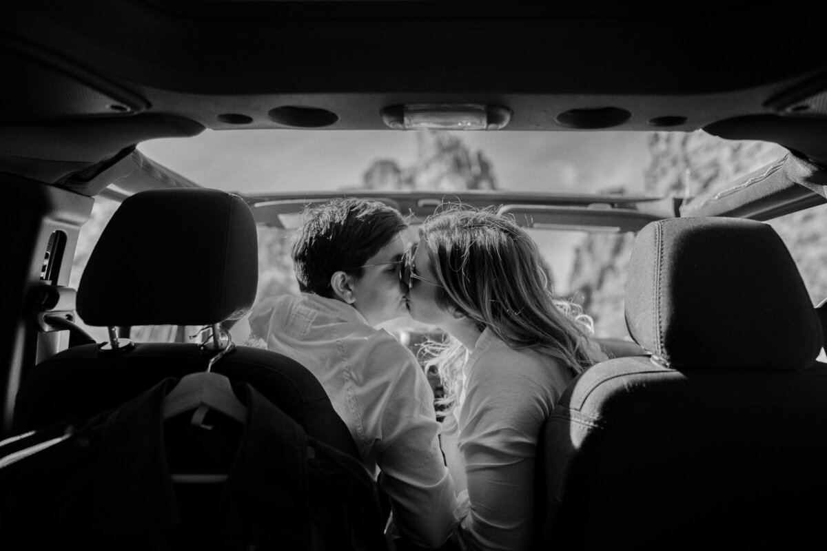 Black and white photo of two brides sitting in the front of a Jeep Wrangler, leaning into the middle for a kiss.