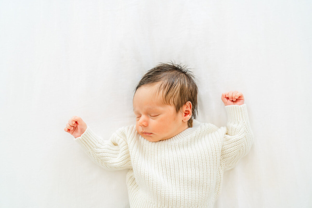 a sleeping newborn baby dressed in an ivory sweater lays on a white sheet