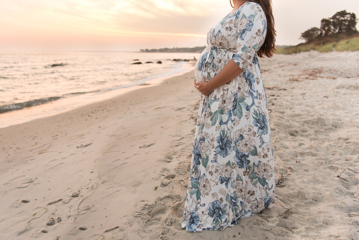Expecting mom is standing by the water in floral dress at sunset, cropped to show just connection to baby