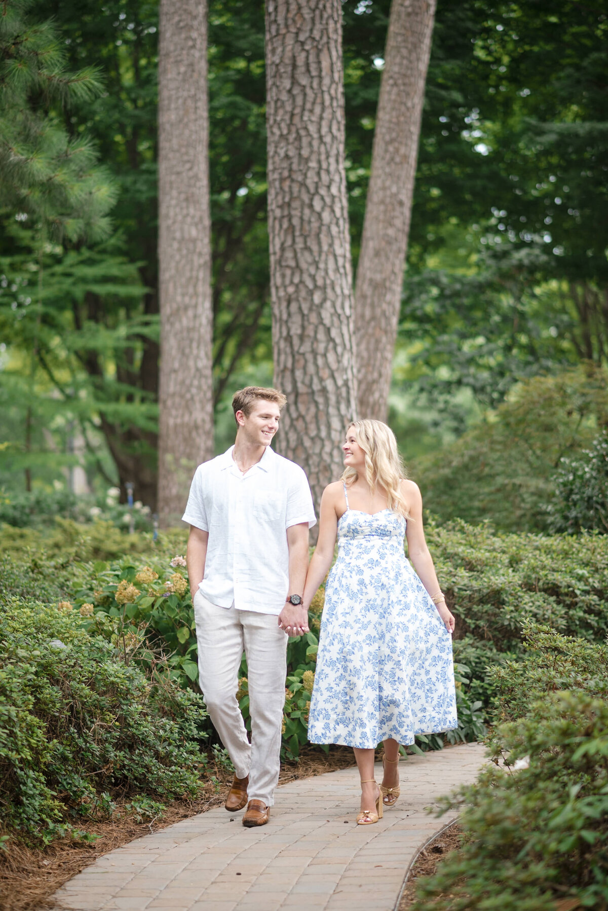 Engaged couple walking hand in hand through a lush garden pathway surrounded by tall trees and greenery in Raleigh, NC—captured by DeLong Photography, a top Raleigh wedding photographer highlighting the most romantic and natural photo locations in the Triangle. 