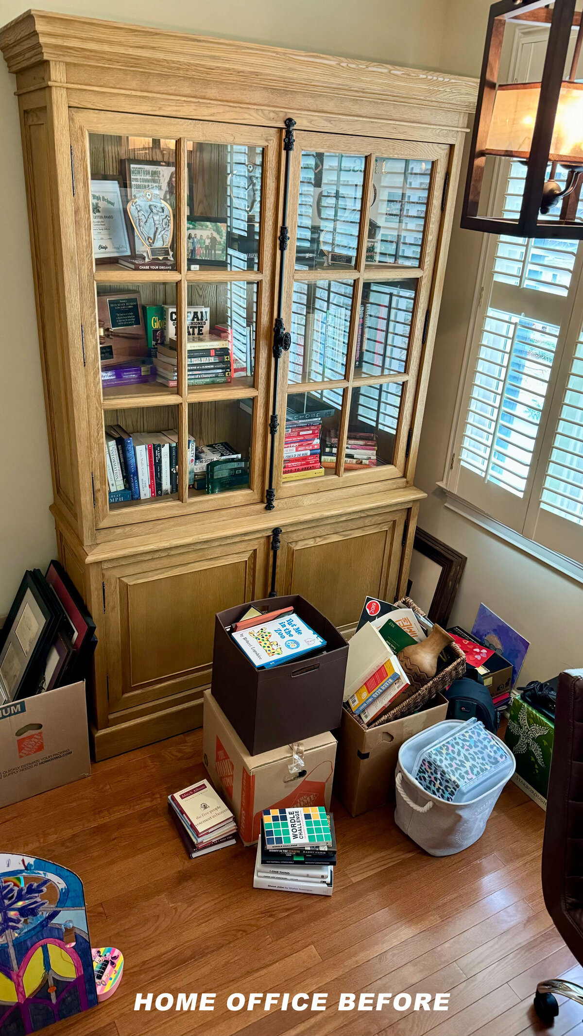 A cozy home office with a wooden bookcase on the left, a leather rolling chair facing a desk on the right, and a patterned rug on a wooden floor. A lantern-style pendant light hangs from the ceiling. Blinds cover the windows.
