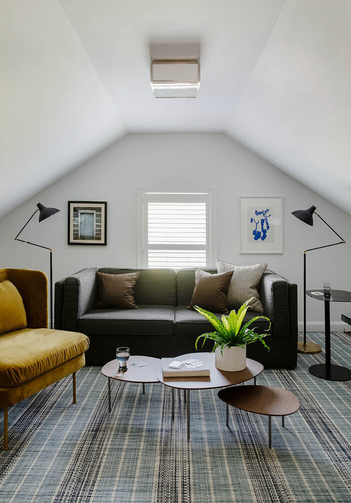 Explore the versatile attic space in a historic Hingham home, serving as an office and guest area with a pull-out sofa, white walls, contemporary nesting tables, and a velvet lounge chair.