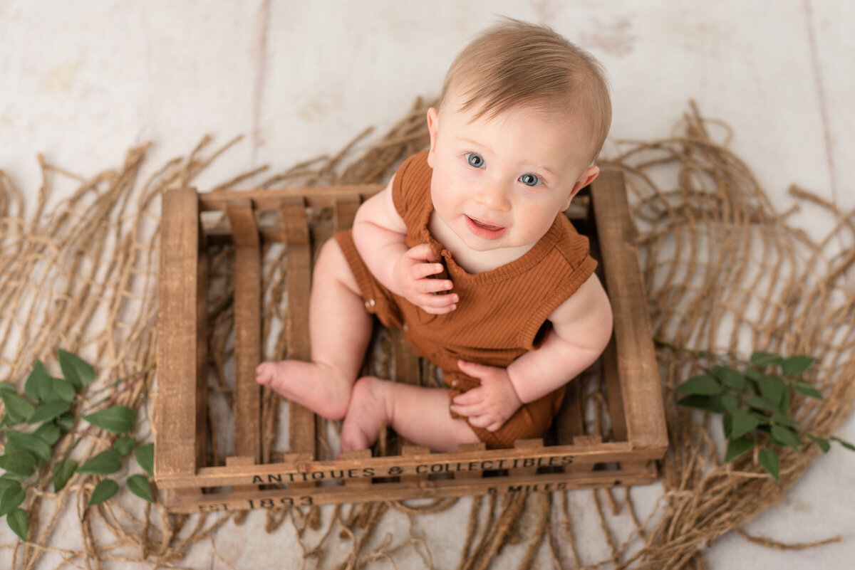 Baby boy smiles up at camera, sitting in crate at rustic session