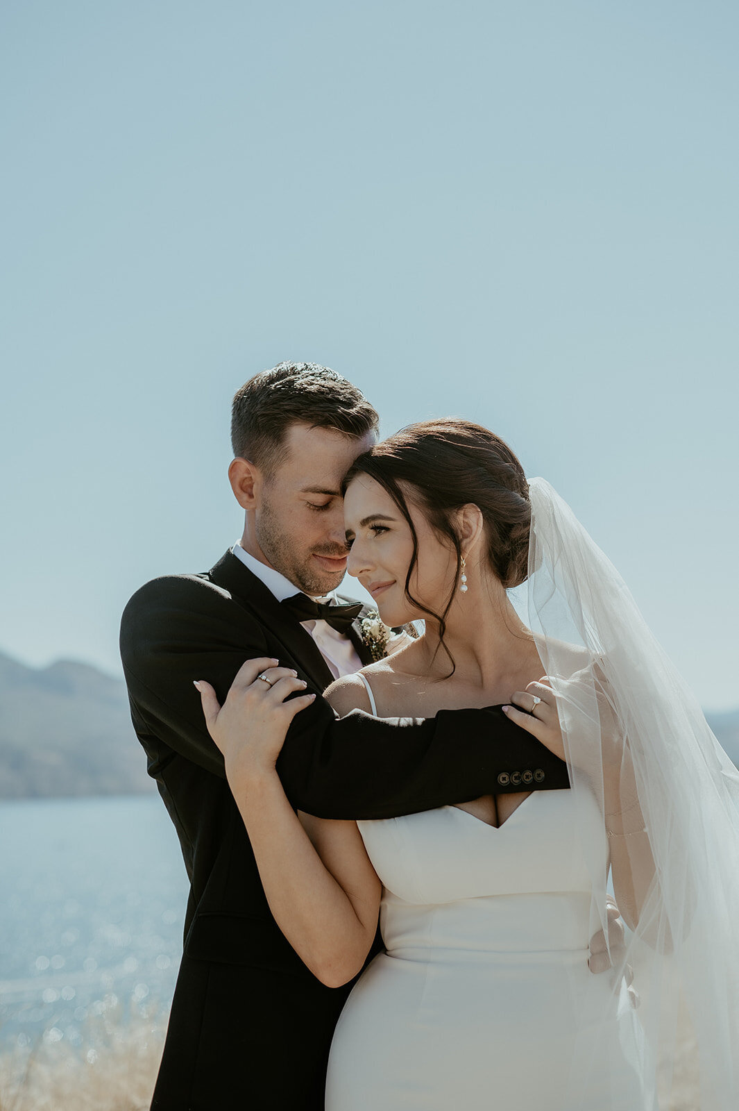 Modern bride and groom in black tux hold each other near a lake.