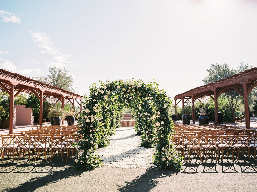 An enchanting wedding ceremony setup featuring a lush floral arch, rustic wooden chairs, and scenic desert views for a dream wedding in Scottsdale.