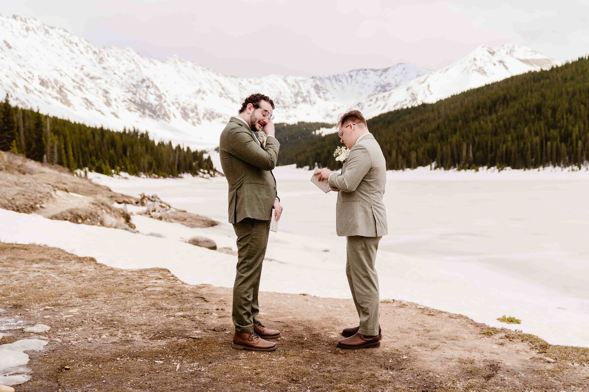  Two grooms exchange emotional vows beside the frozen Clinton Gulch Dam Reservoir in Colorado, surrounded by snow-capped mountains and evergreen forests.