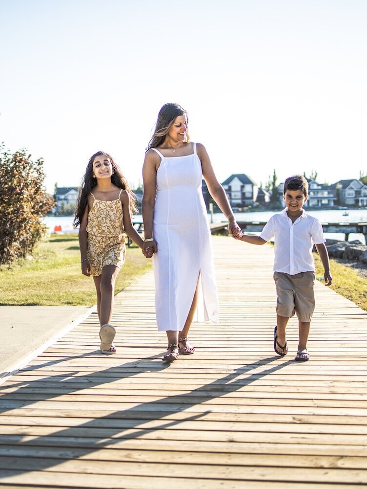 A mom in a white dress is walking near a lake holding her son and daughter's hands