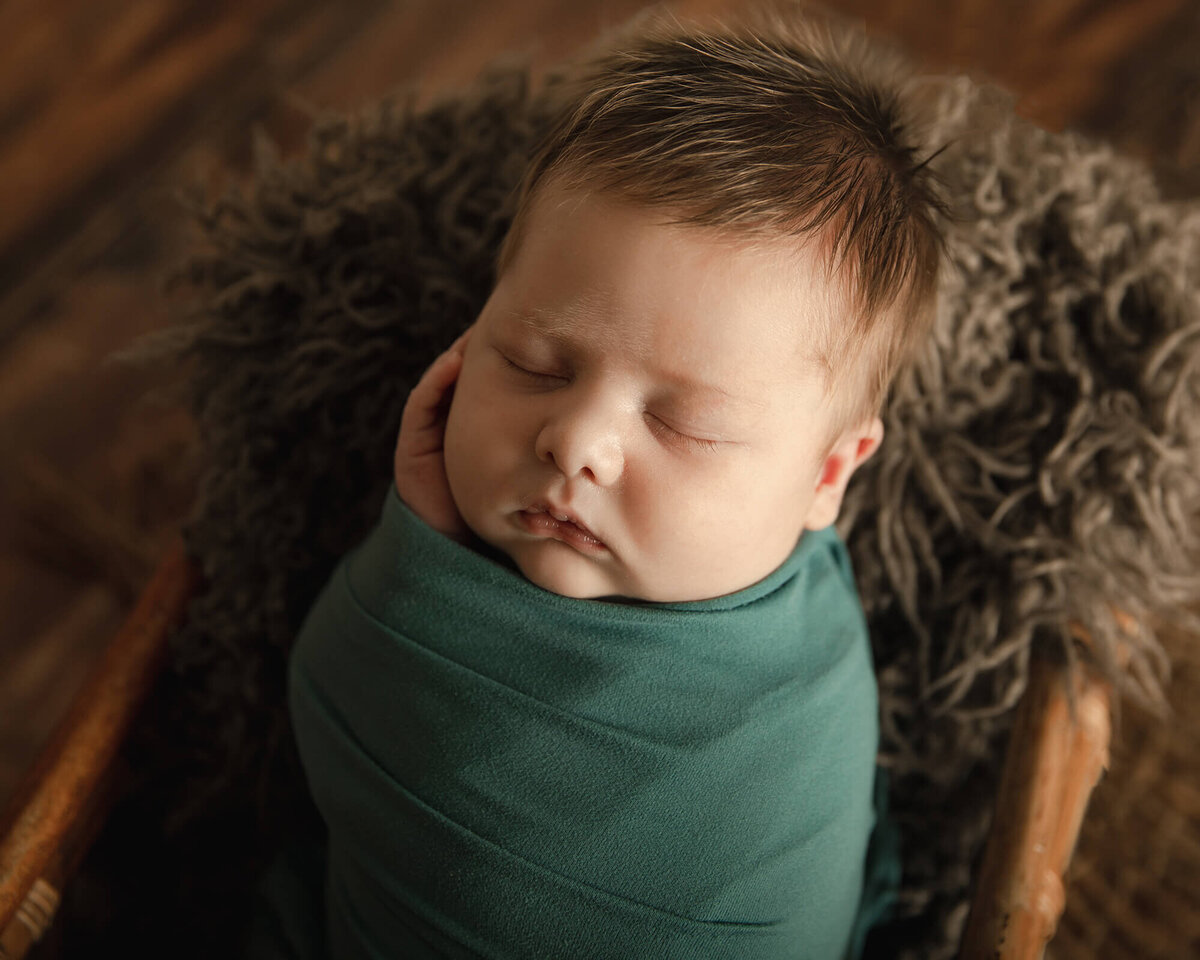 Newborn boy with lots of light brown hair laying in basket with his hand cupping his face. He is wearing a teal wrap.