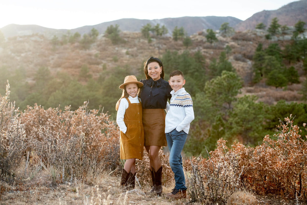 A mother in a black top and brown skirt stands on a trail with arms around her son and daughter