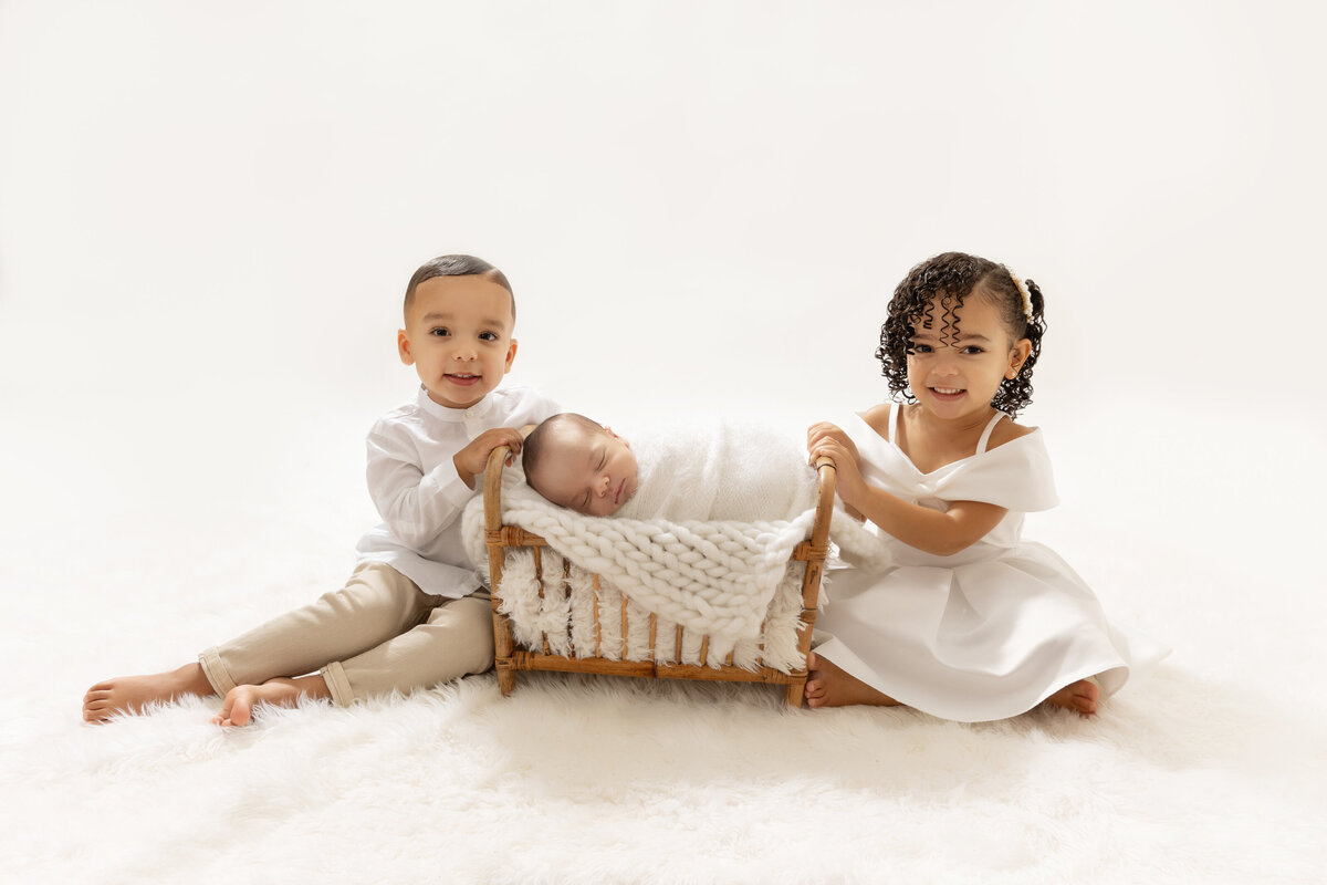 Happy toddler brother and sister in white outfits sit on the floor of a studio with their sleeping newborn sibling in a crib between them