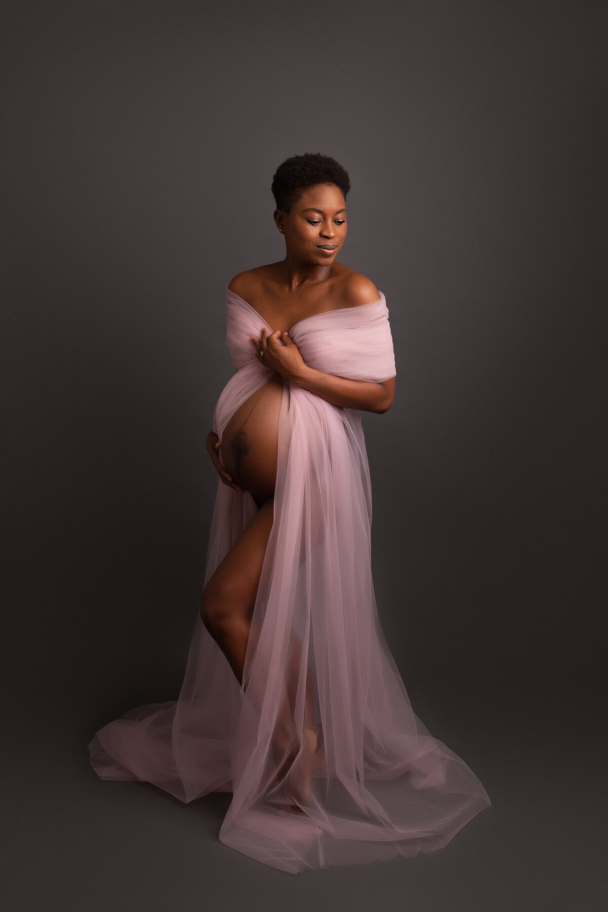 London studio maternity portrait of a woman draped in pink tulle fabric on a grey background.