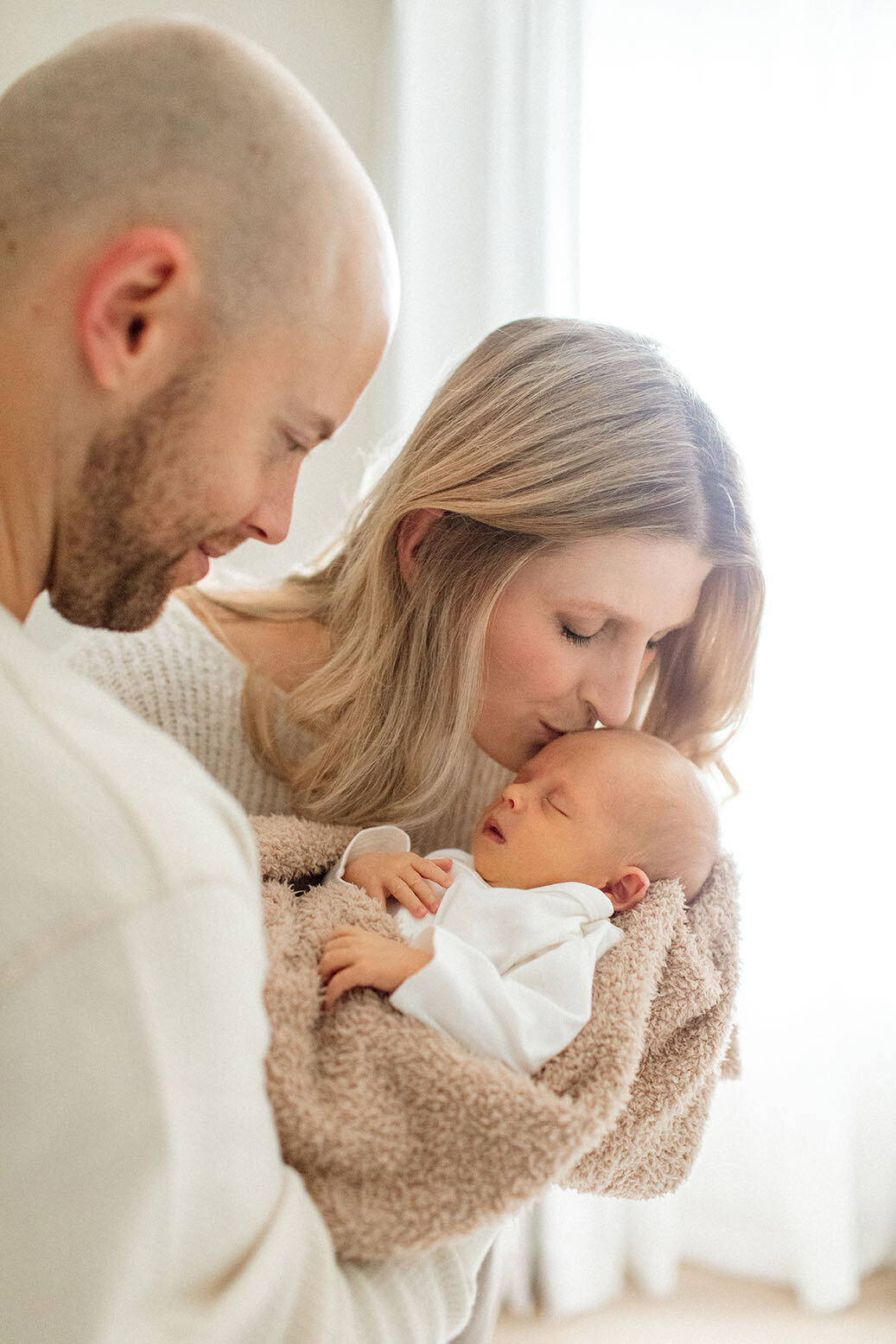 Parents kissing newborn baby together - Sara James documents tender new parent moments and first family bonds during intimate newborn photography sessions in San Antonio Texas homes
