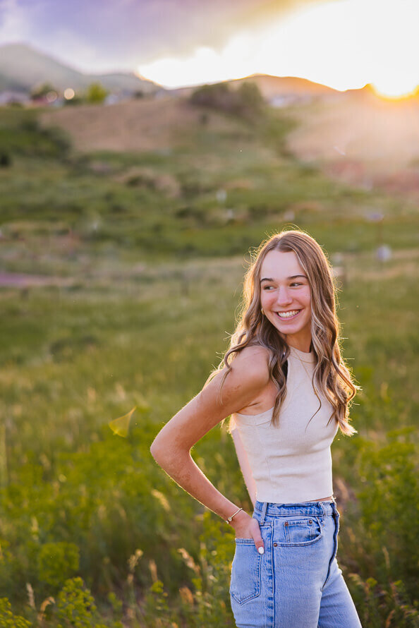 Senior girl puts hands in back pockets and looks over her shoulder in a grassy field.