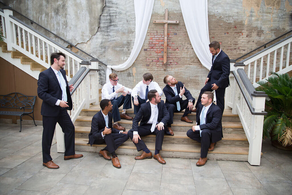Seven men in suits sit and stand casually on wooden stairs, talking and laughing together. Behind them is a large cross, white drapery, and a weathered brick wall, giving a relaxed, rustic atmosphere.