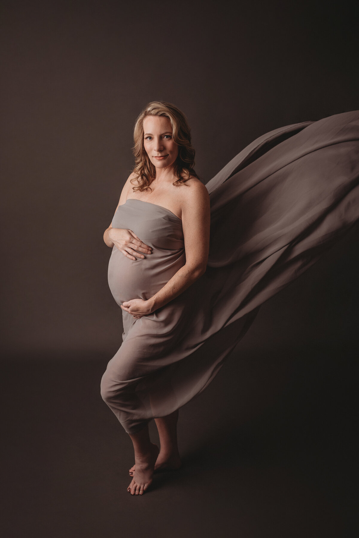Pregnant woman photographed by atlanta maternity photographer Casey McMinn Photography standing on gray backdrop draped in gray chiffon fabric holding baby bump looking at camera