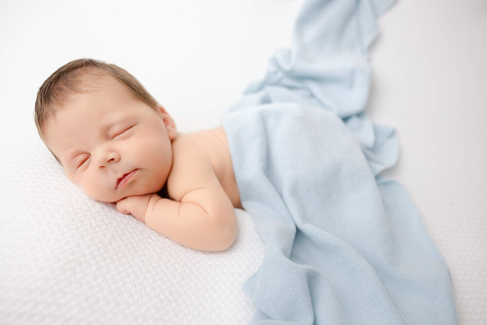 Sleeping baby boy with hand under chin and baby blue blanket covering body.
