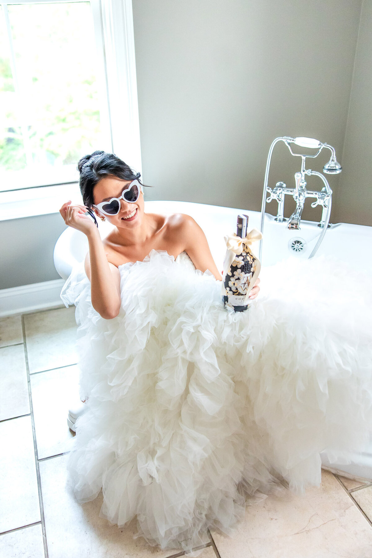 Bride playfully posing in a bathtub in her wedding dress, holding a bottle of champage
