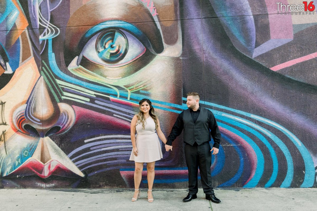 Soon to be Groom holds his fiance's hand and looks at her in front of a large mural