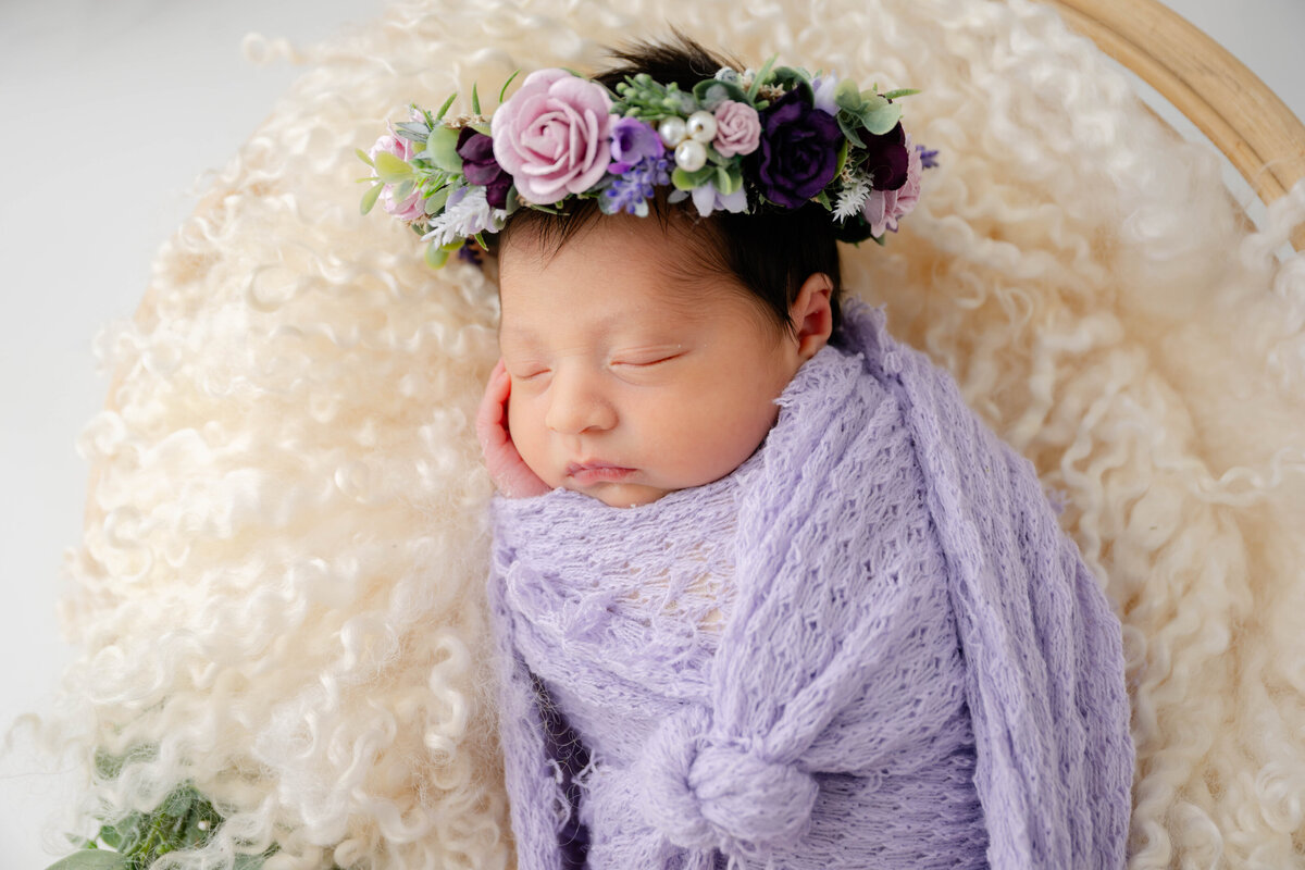 Close up of a baby with hand on cheek while swaddled in a lilac wrap and a flower crown on her head. 