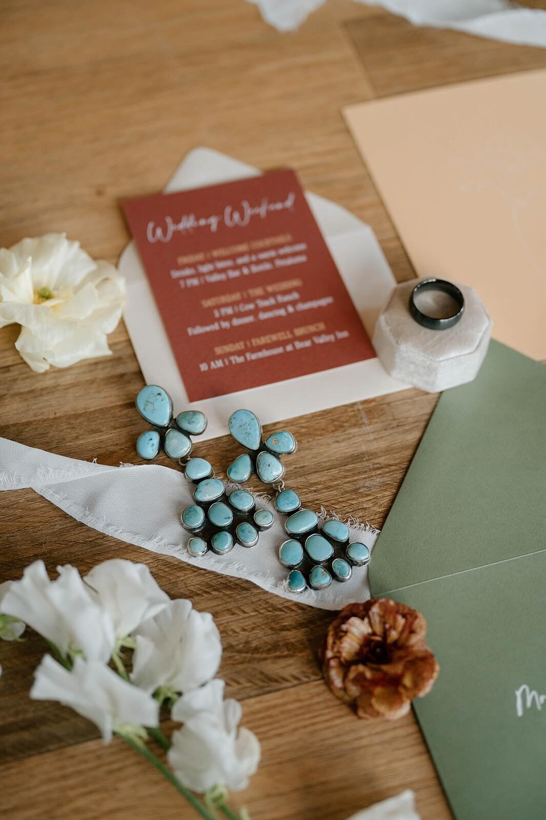 Wedding invitation flatlay with terracotta card, white ribbon, turquoise earrings, and flowers.