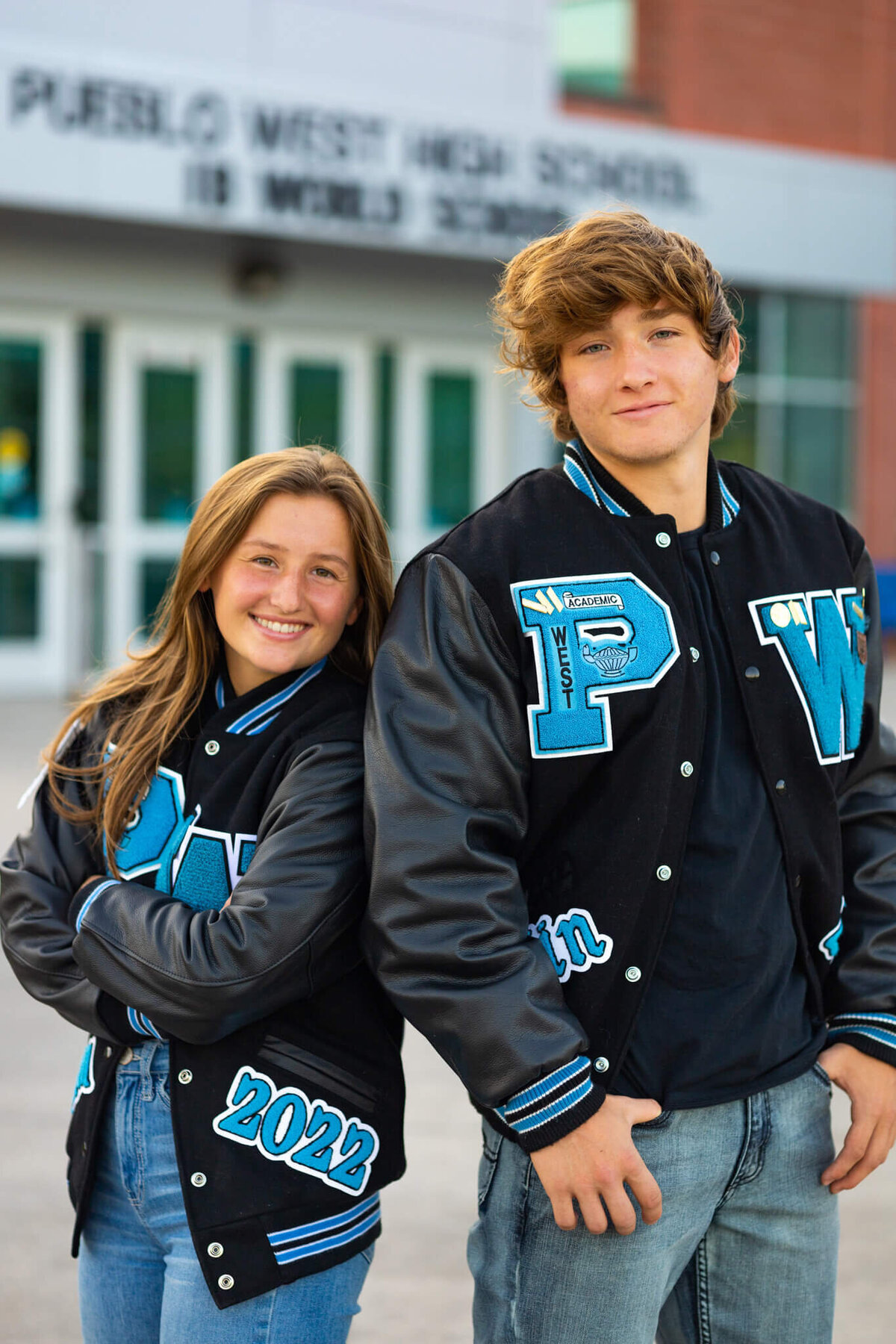 twin brother and sister wearing their letterman jackets standing in front of their high school