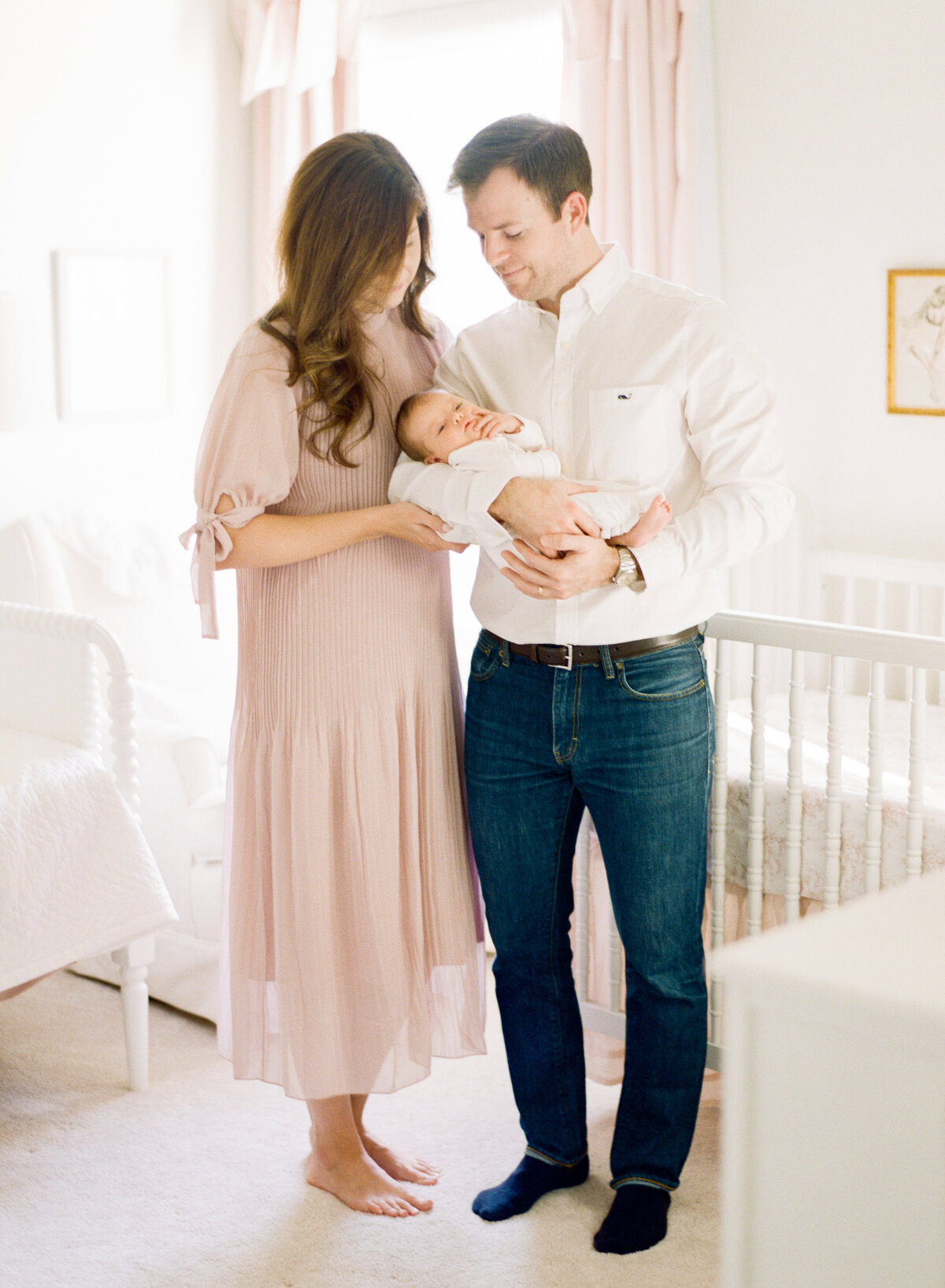 New parents hold their baby and stand in front of her crib. Image by Raleigh newborn photographer A.J. Dunlap Photography.