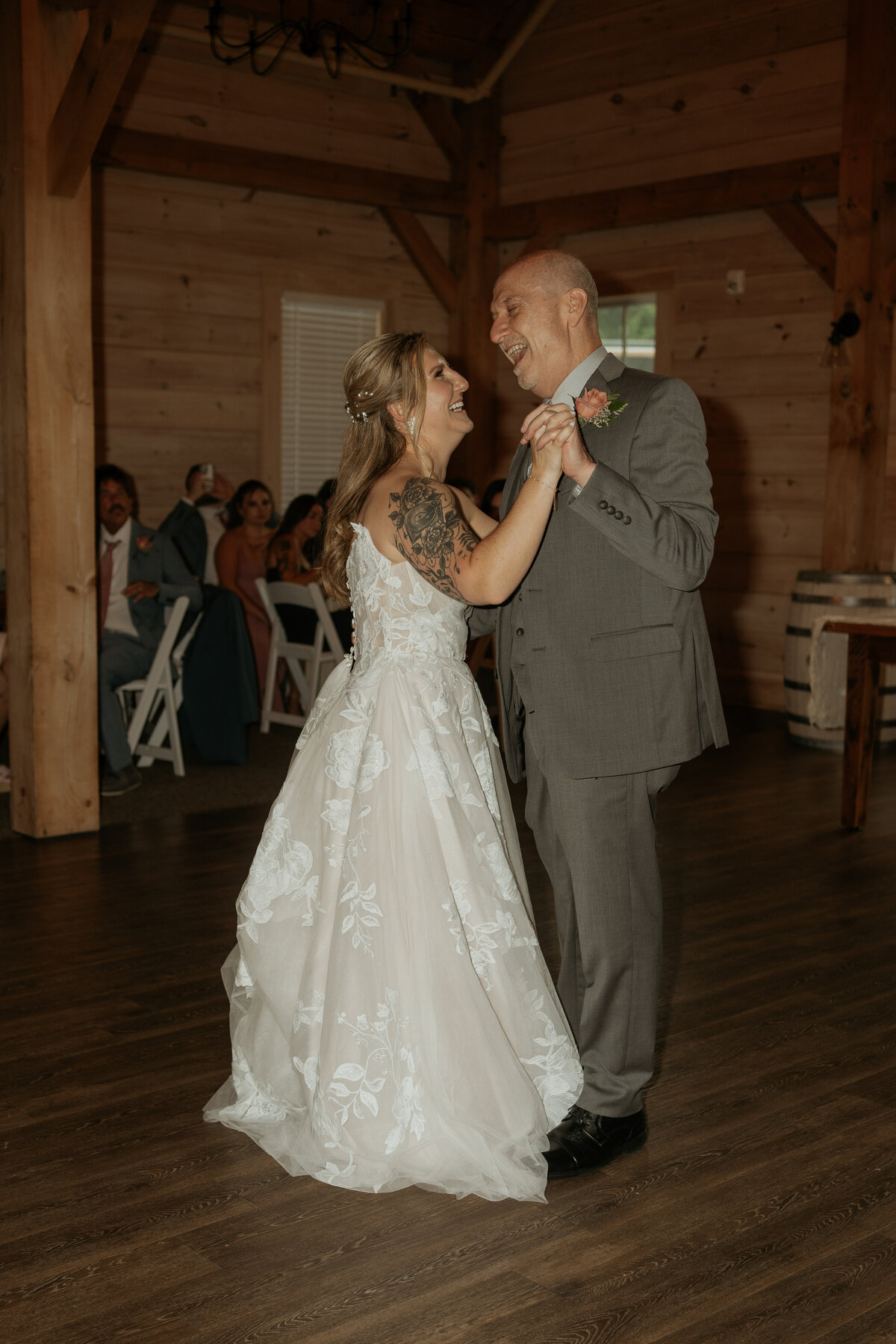 Bride and her father share a joyful dance at a rustic indoor wedding venue. The bride, in a lace gown with floral details and a visible arm tattoo, and her father, in a grey suit, smile and laugh together. The warm wooden interior and the presence of guests in the background add to the intimate and celebratory atmosphere.