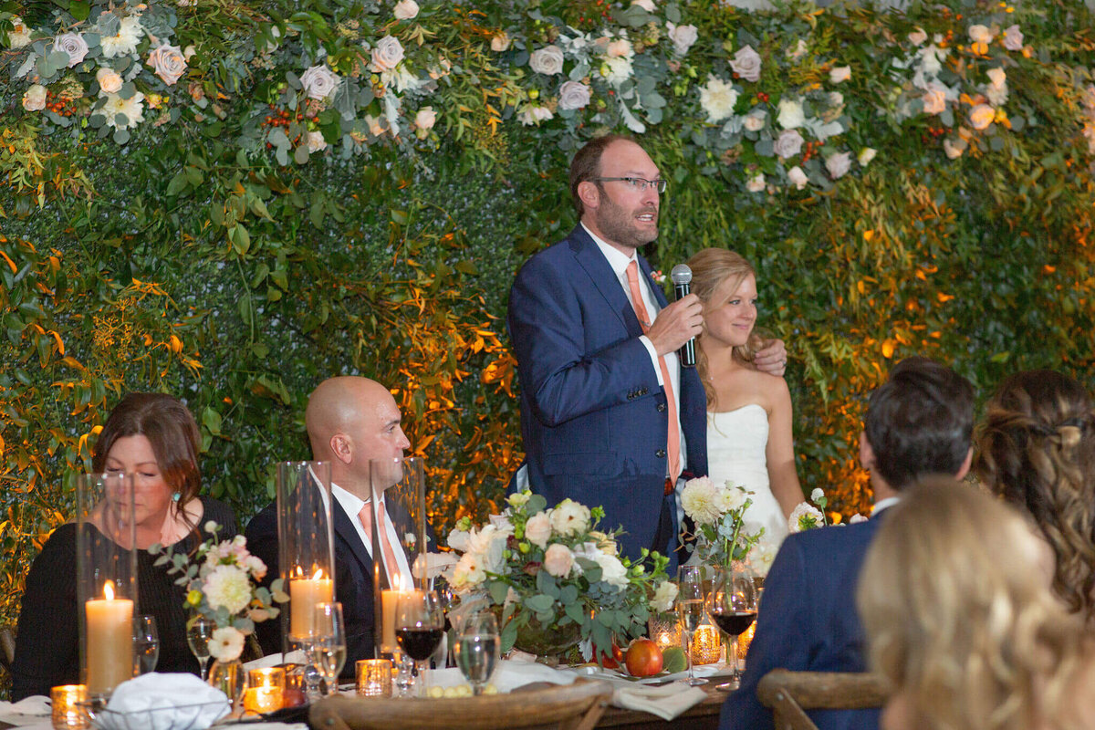 bride and groom stand to deliver a toast, surrounded by their wedding party