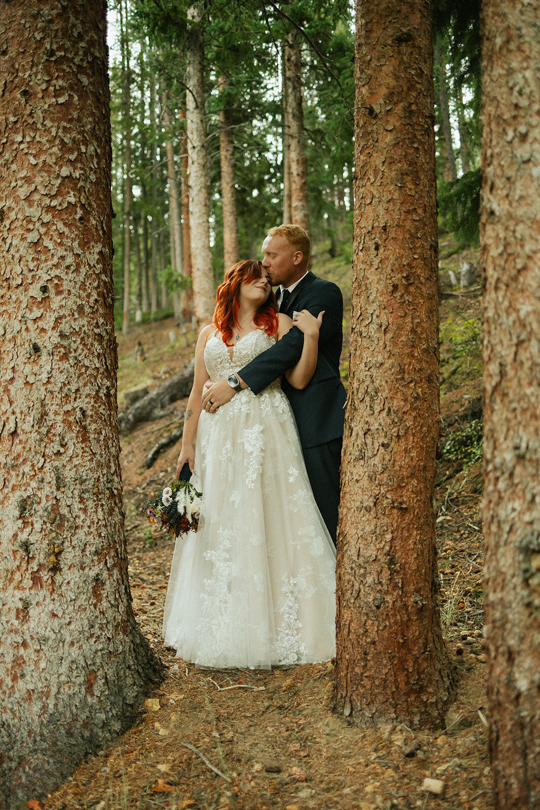 Groom kissing brides forhead with arm across standing in forest