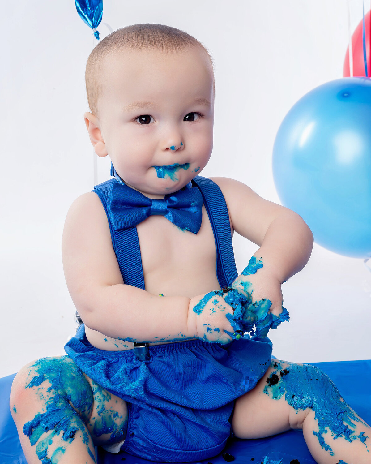 Baby in a blue outfit with blue frosting on hands and legs, with balloons in the background.