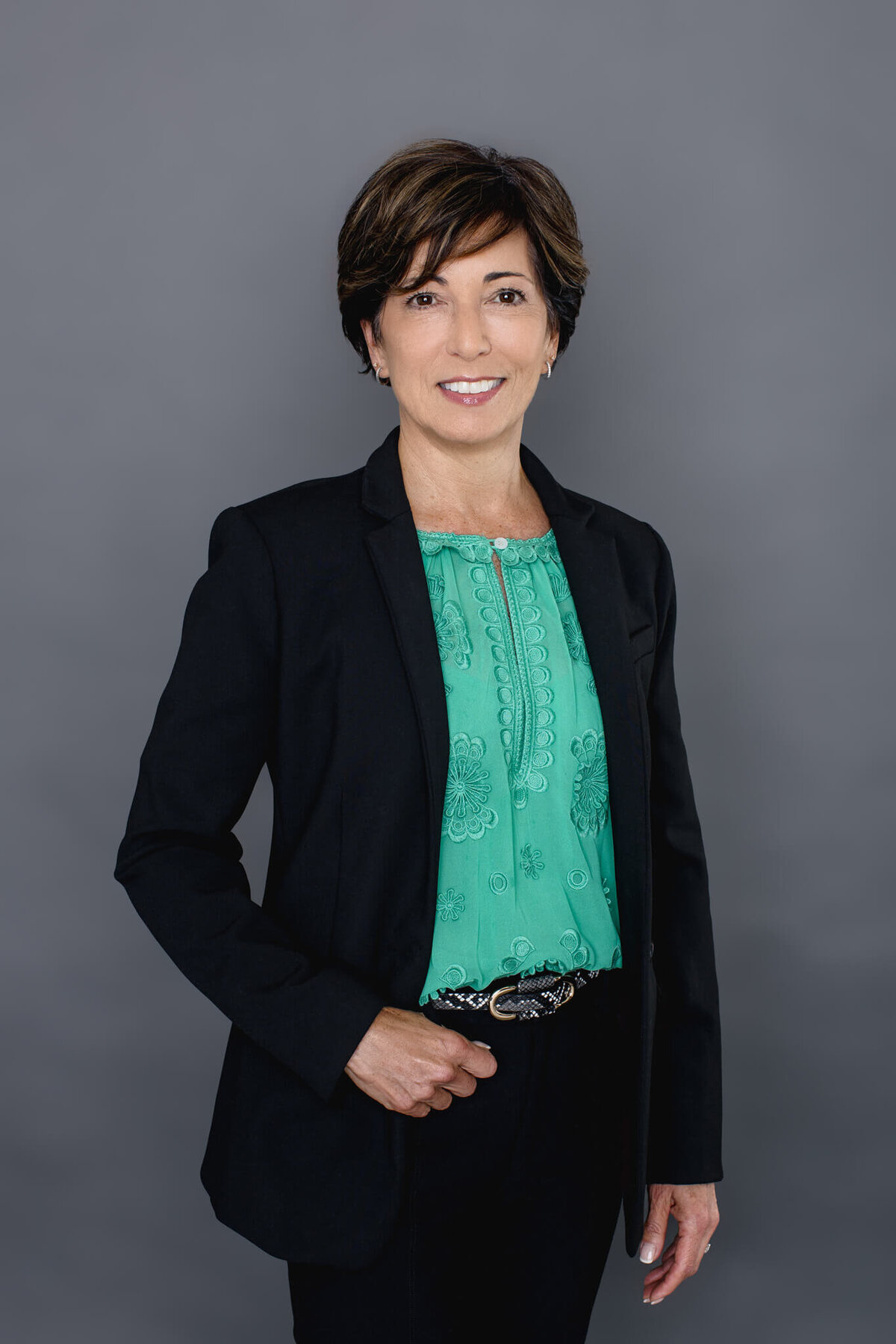 An older woman with short hair wearing a light green blouse and a black blazer, smiling at the camera with her right hand in the belt loop of her black pants in front of a grey background at a studio in Tustin.