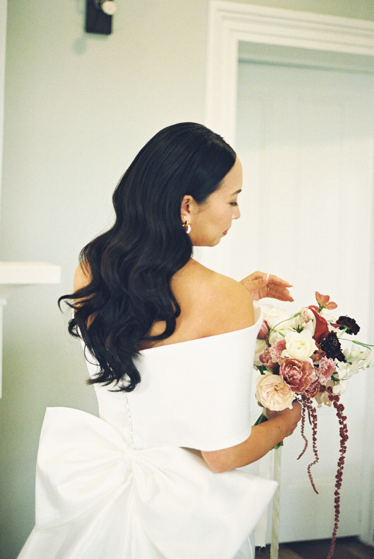A bride with long hair in an off-shoulder white gown with a large bow on the back holds a bouquet of roses and flowers.