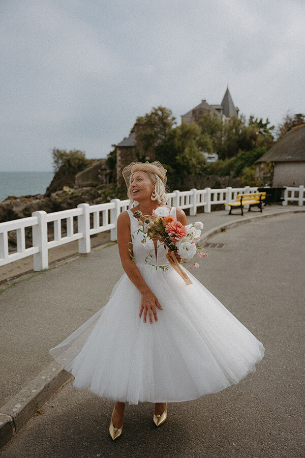 La mariée fait tournoyer sa robe, bouquet en main, sur la route. Instant de bonheur en bord de mer en Bretagne, par Kamescop.