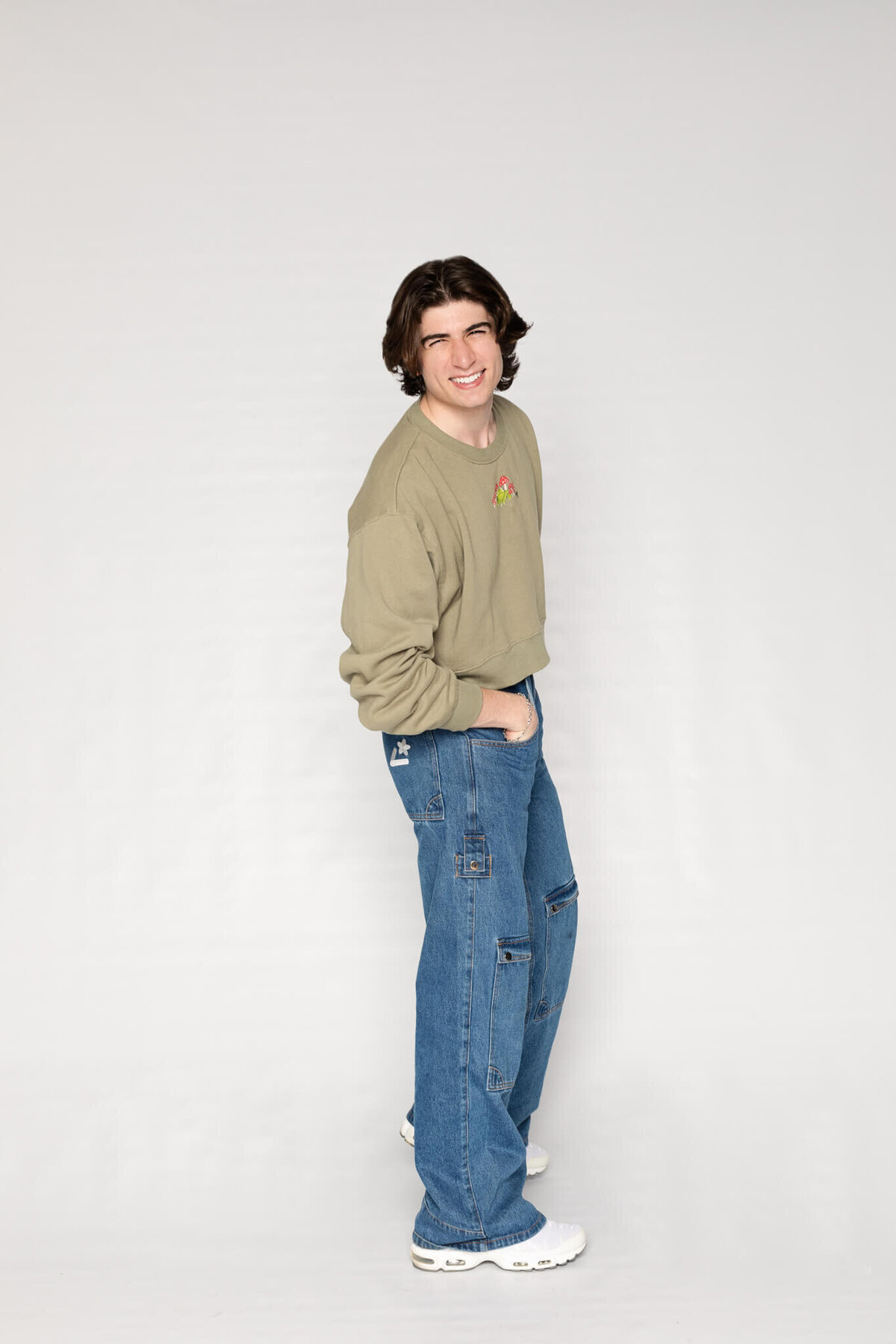 A person with shoulder-length hair smiling, wearing a khaki sweater and blue jeans with white sneakers. They stand against a plain gray background, with one hand in their pocket and a relaxed posture.