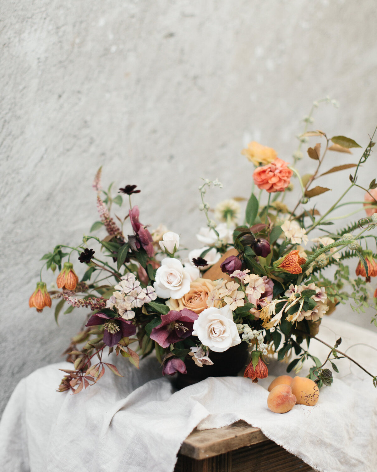 An arrangement of organic, purple and orange summer flowers sits on a tablecloth on a wood table.