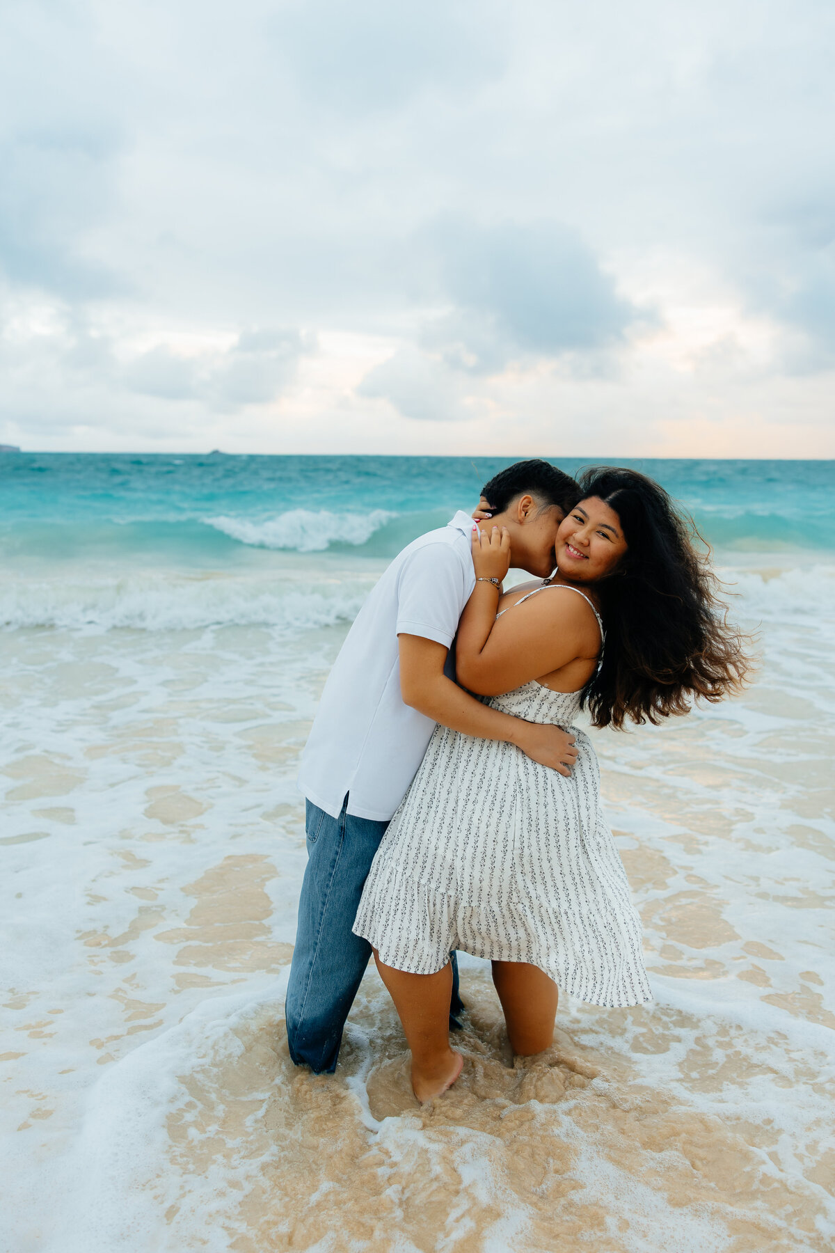 Couple embracing by the shoreline as waves crash behind them during their Oʻahu couples photoshoot