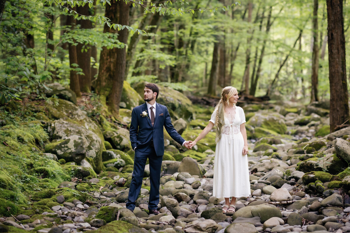 bride and groom holding hands and looking opposite directions in a river rock bed in the woods near Spence Cabin