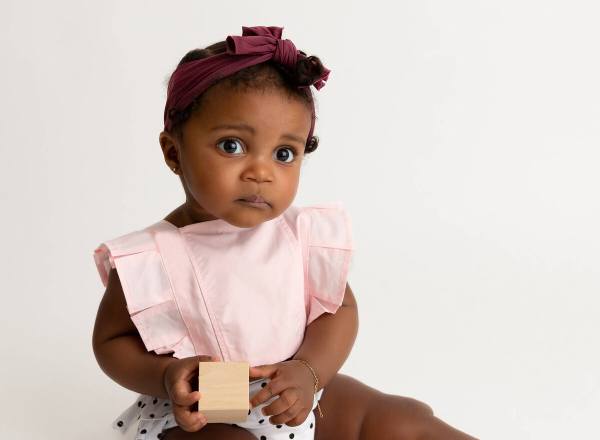 6-month-old baby girl sitting and smiling at the camera while holding a wooden block during a milestone photoshoot by NYC baby photographer