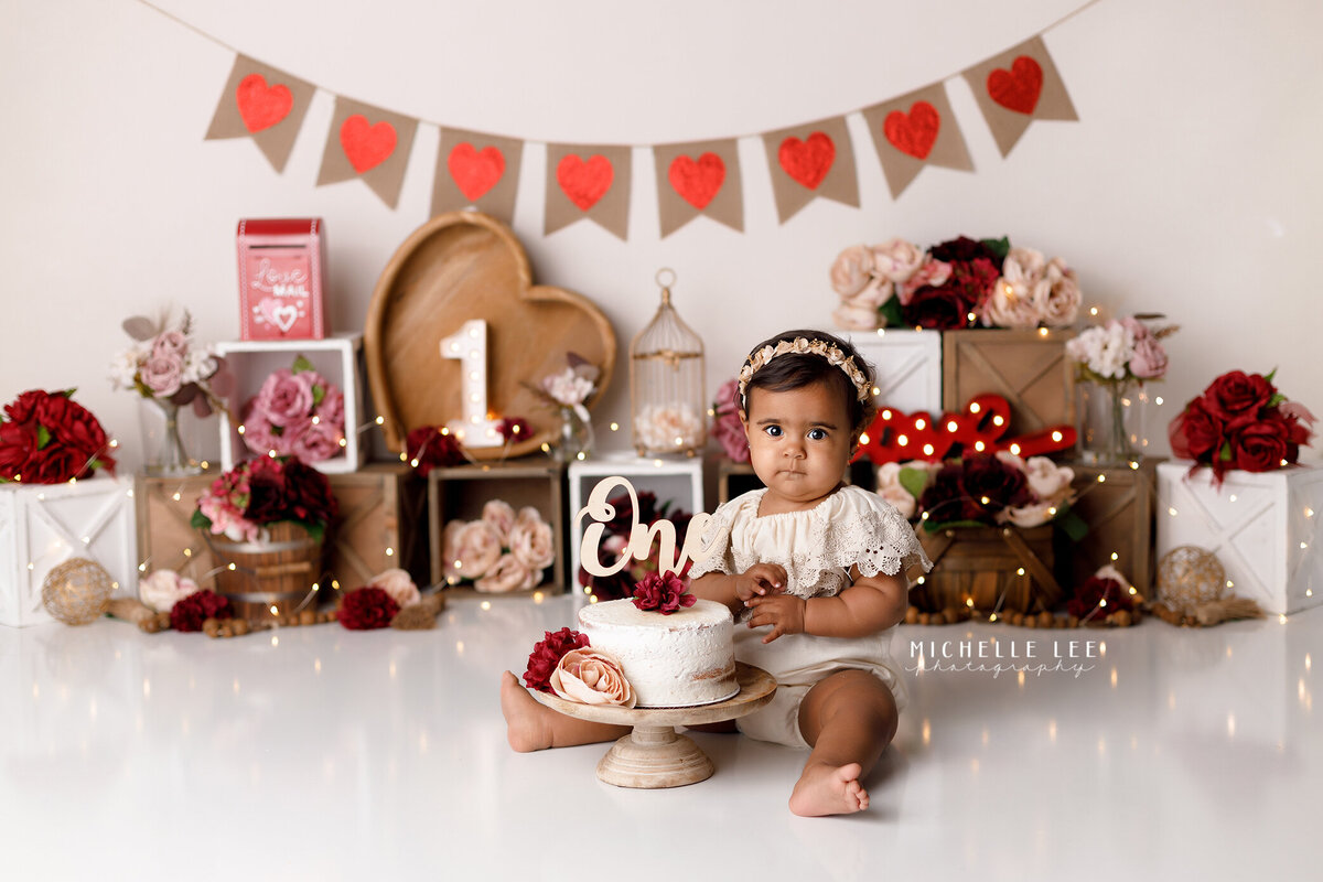 Rustic Valentine's Day themed cake smash in Palm Beach and Boynton Beach photography studio. Baby girl is wearing a BoHo off the shoulder romper sitting behind a white floral decorated cake. In the background is various valentines day decorations with red, pink and maroon flowers.