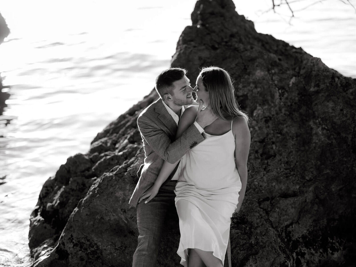 Black and white photo of the engaged couple sitting on a big rock, sweetly staring at each other at the seashore in Jamaica.