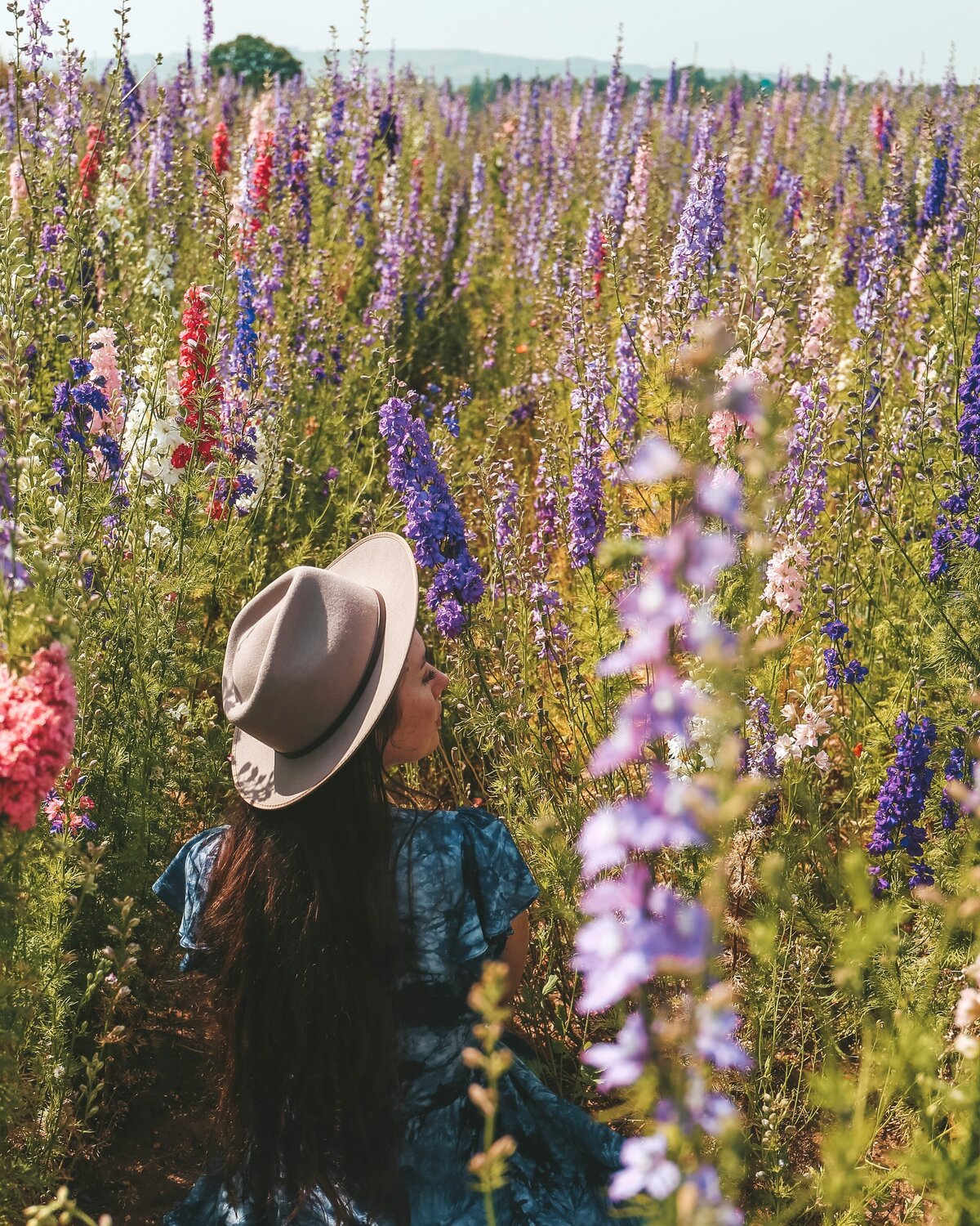 A young woman sits in a sunlit field of wildflowers, her flowing dress blending with the soft hues of nature. Surrounded by delicate blooms, she embodies cottagecore charm and a dreamy, storybook-inspired escape.