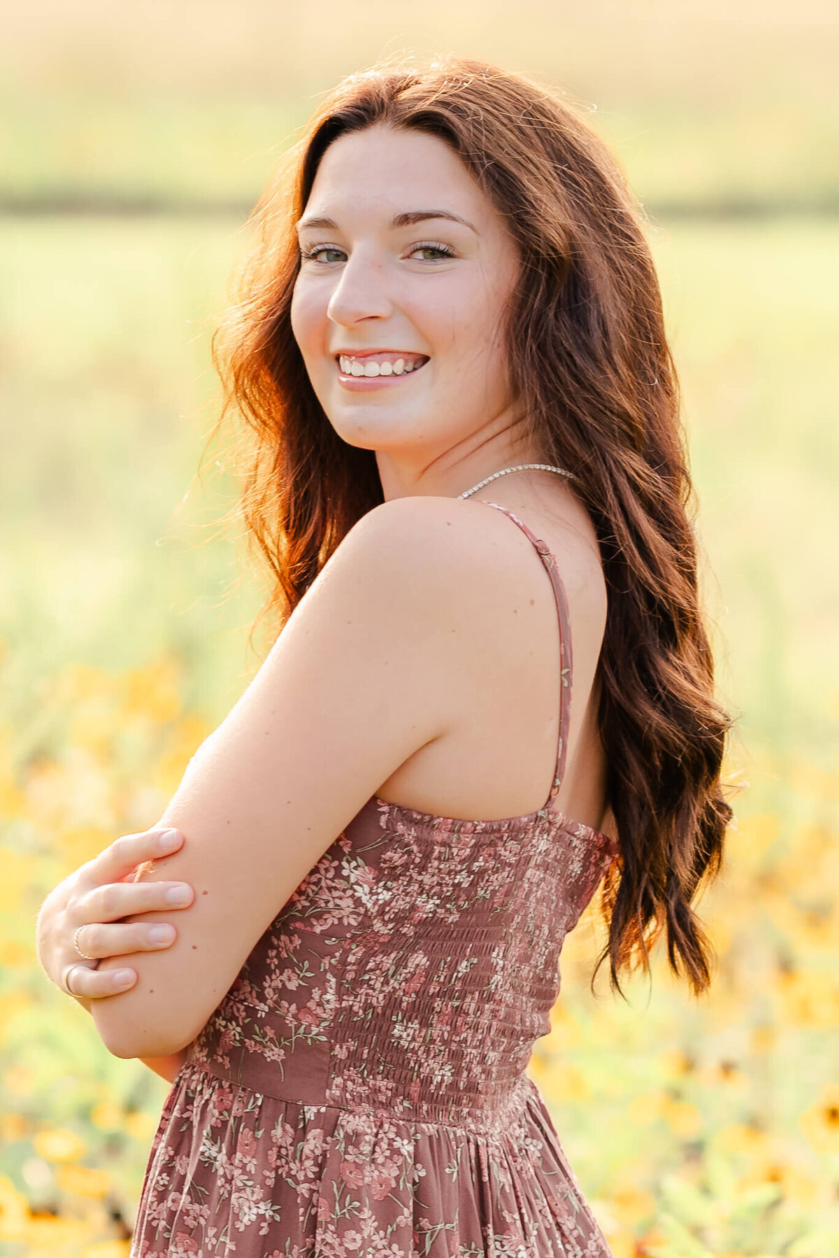 A senior wearing a floral dress and standing in a field of yellow flowers crosses her arms and looks over her shoulder, smiling.