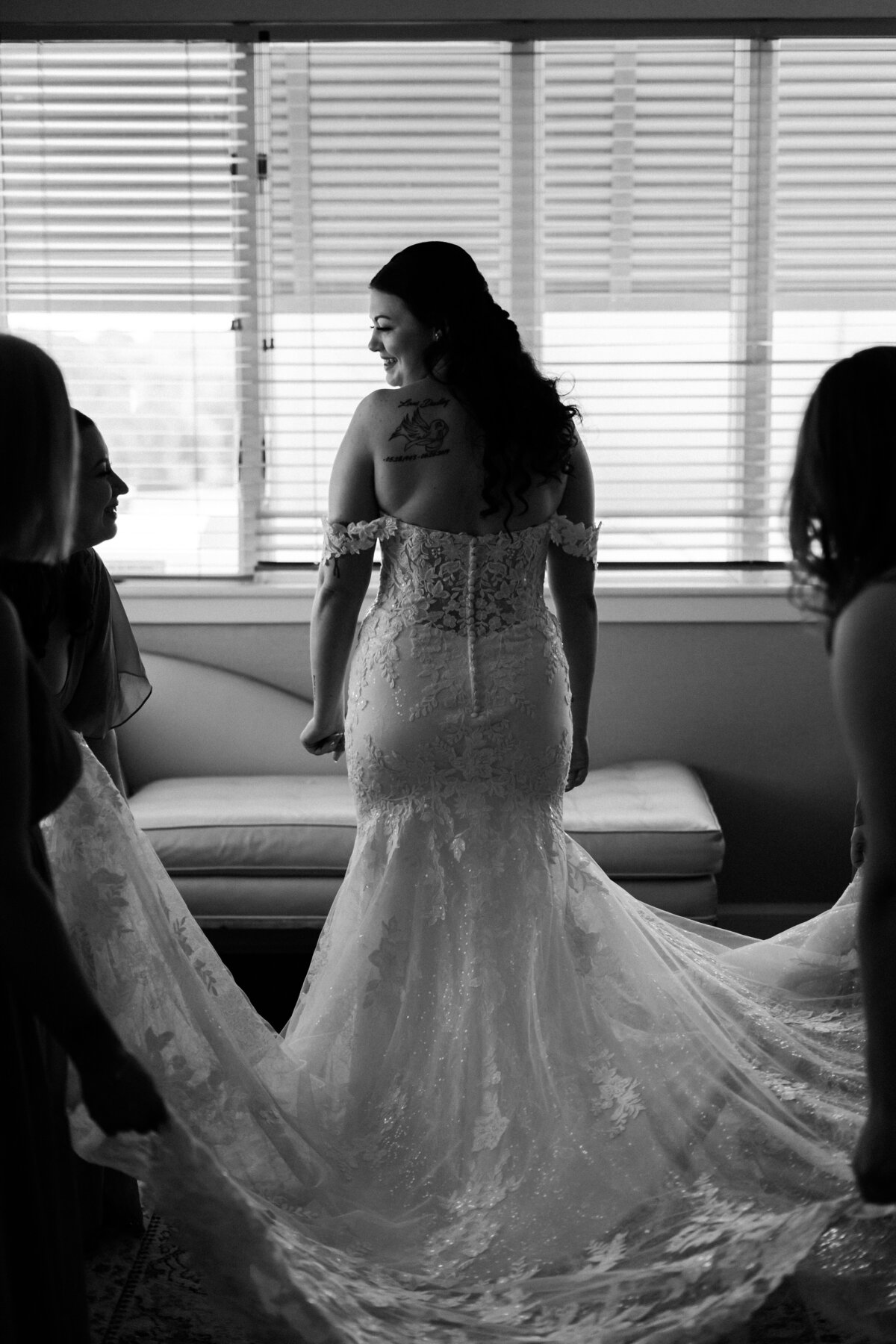 A beautiful bride in a detailed off-the-shoulder lace wedding gown is captured from behind, looking over her shoulder with a subtle smile, as she prepares for her wedding at the Lesner Inn in Virginia Beach, photographed by Quinn Photography LLC.